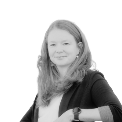 Black-and-white portrait of a woman with long, light-colored hair, wearing a dark cardigan and earrings, smiling gently at the camera. She wears a smartwatch on her wrist.