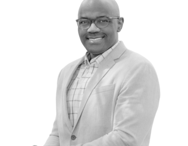 Black-and-white portrait of a smiling bald, Black, man wearing rectangular glasses, a light-colored herringbone blazer over a windowpane-check collared shirt, and a wristwatch. He stands at an angle with one hand resting on the other, set against a plain white background