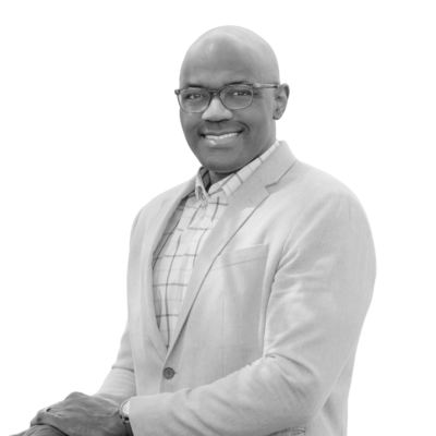 Black-and-white portrait of a smiling bald, Black, man wearing rectangular glasses, a light-colored herringbone blazer over a windowpane-check collared shirt, and a wristwatch. He stands at an angle with one hand resting on the other, set against a plain white background