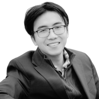 Smiling Asian man with dark hair and rectangular glasses, wearing a dark blazer over a zip-neck sweater, in a black-and-white headshot.