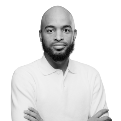 Black-and-white portrait of a bald Black man with a full beard, wearing a light-colored polo shirt and looking directly at the camera with his arms crossed.